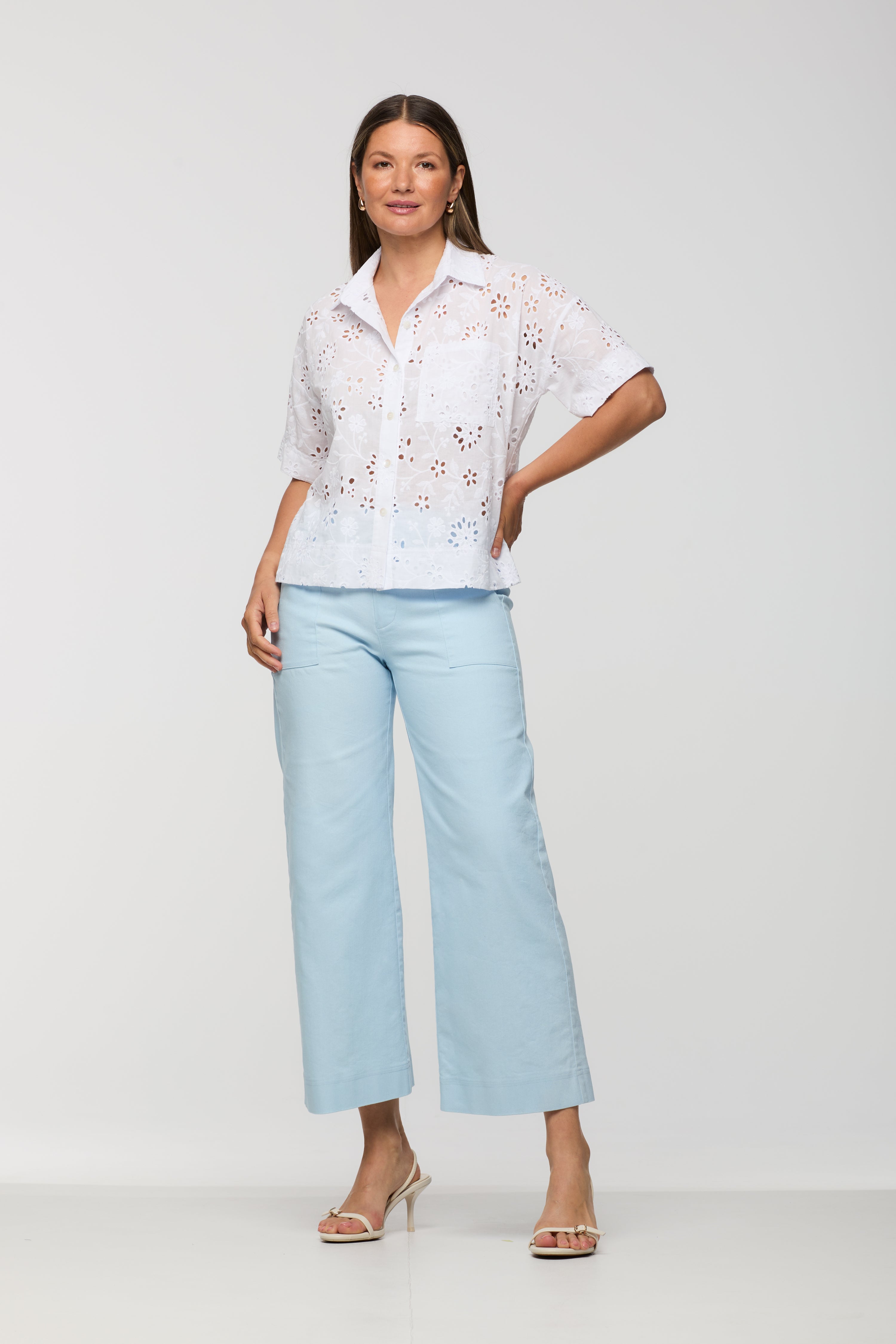 Woman wearing a short sleeve white embroidered eyelet button-front blouse with a collared neckline and relaxed fit, paired with light blue cropped wide leg pants.