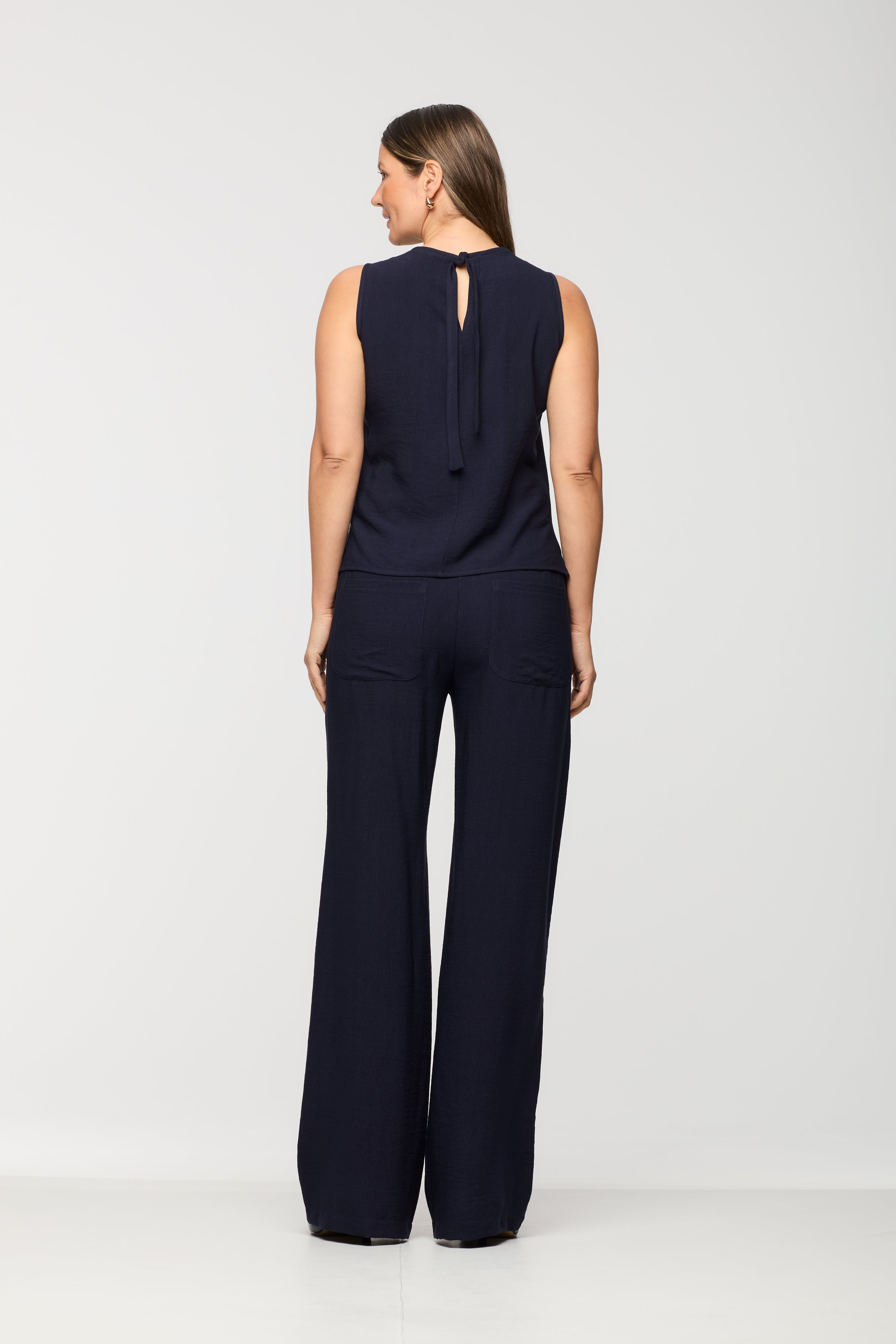 Woman wearing a sleeveless navy top shown from the back, featuring a keyhole neckline with tie detail, paired with matching navy wide leg pants.