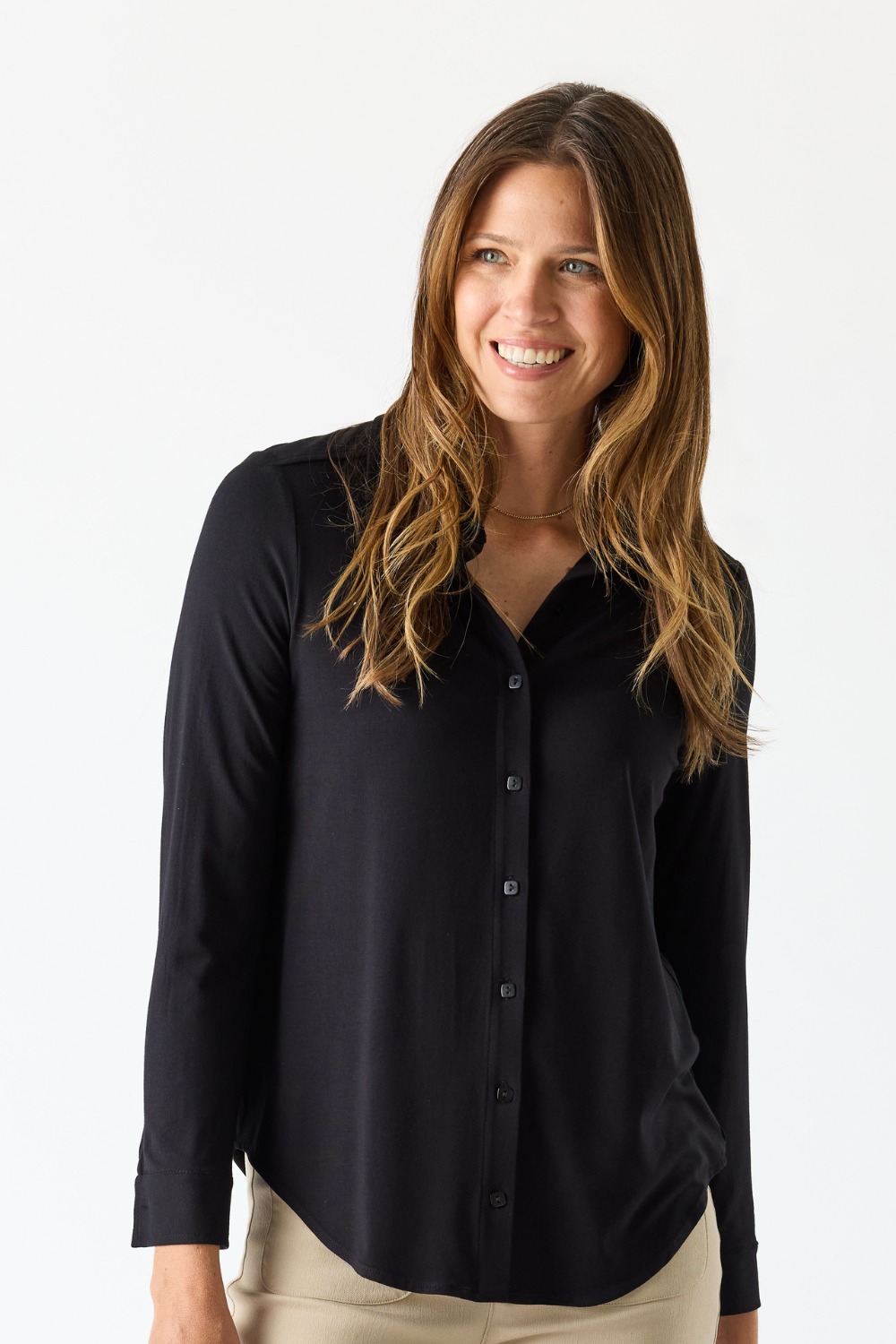 A woman with long brown hair wearing a black long-sleeve button-down shirt and beige pants, photographed against a plain white studio background.
