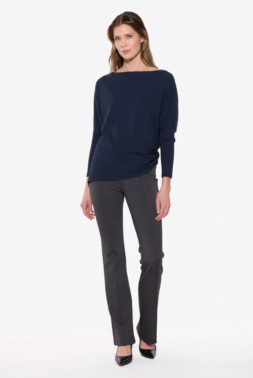 A woman modeling a navy blue boat-neck knit top with long sleeves, paired with dark gray trousers and black heels, against a plain white studio background.