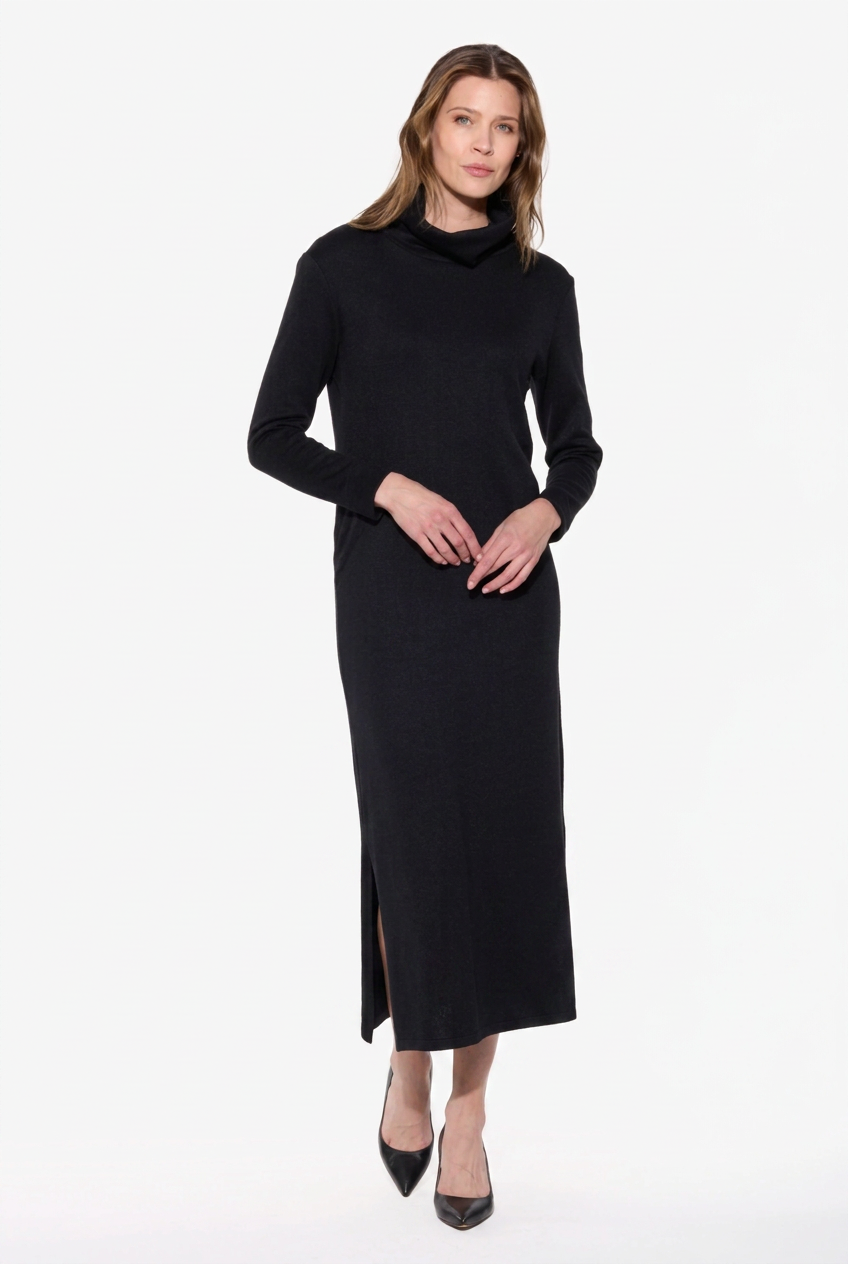 A woman modeling a long-sleeve black mock neck midi dress with a side slit, paired with black pointed-toe heels, against a plain white studio background.