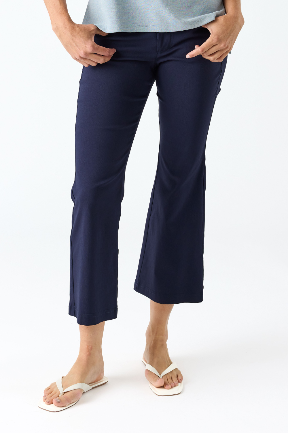 A person wearing navy blue cropped flare pants with hands in pockets, paired with a light blue top and white flip-flop sandals, against a plain white studio background.
