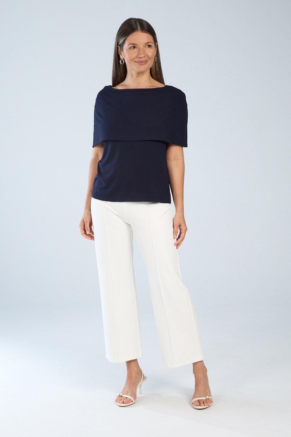 A woman wearing a navy blue top with a boat neckline and short sleeves, paired with white wide-leg pants, standing against a neutral background.