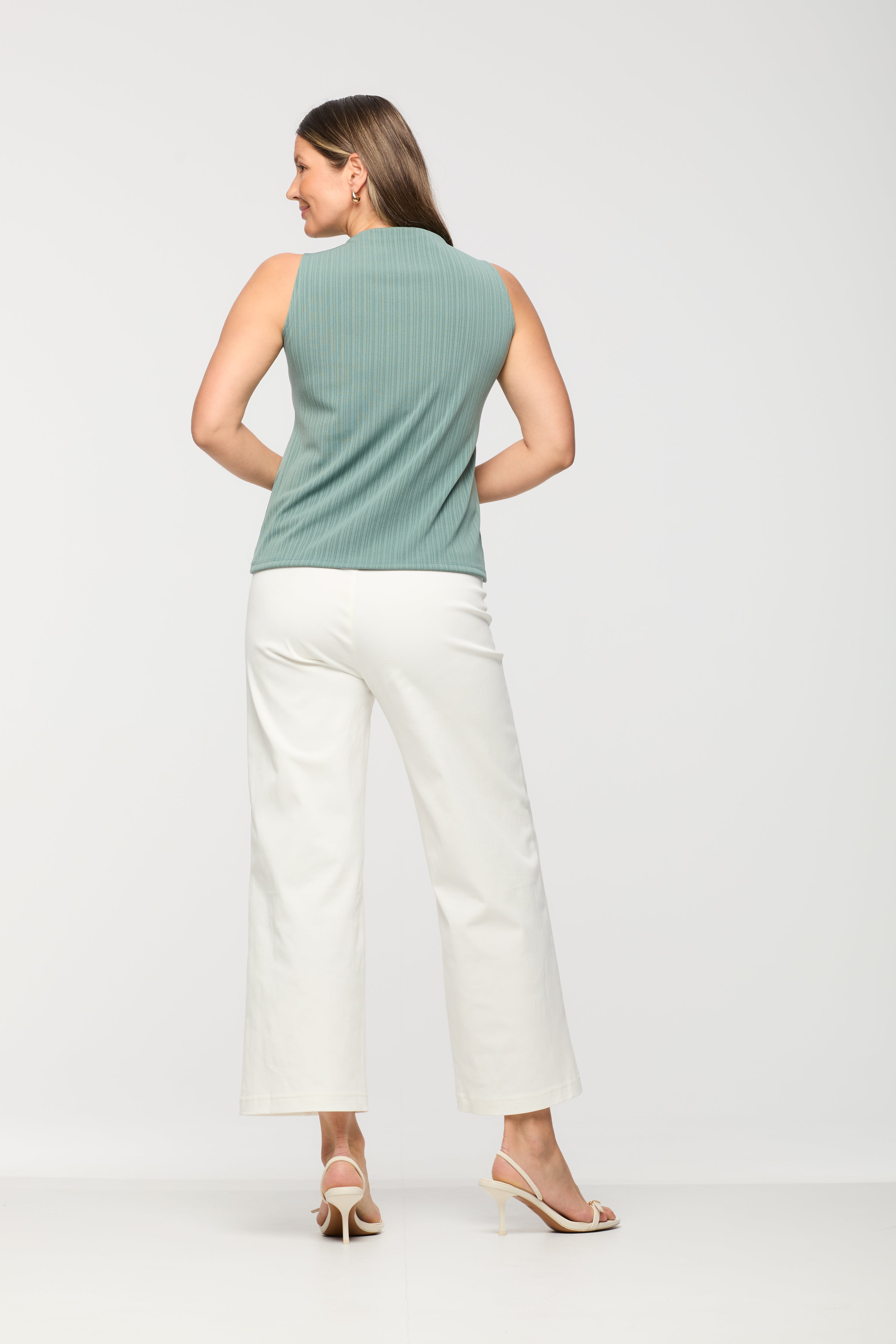 Woman wearing a sleeveless sage green ribbed knit top shown from the back, featuring a high neckline and straight hem, paired with white wide leg cropped pants.