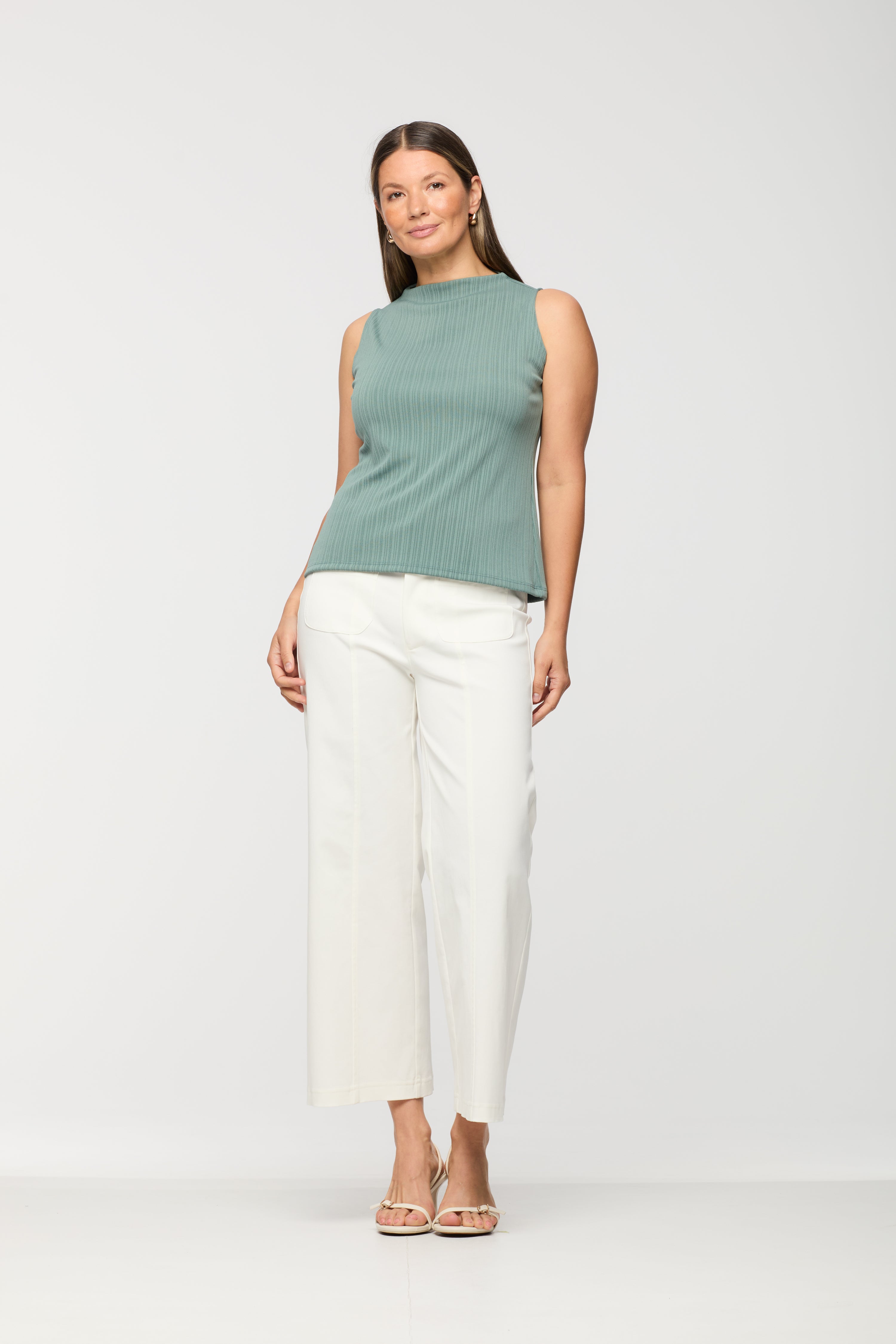 Woman wearing a sleeveless sage green ribbed knit top with a high neckline and relaxed fit, paired with white wide leg cropped pants.