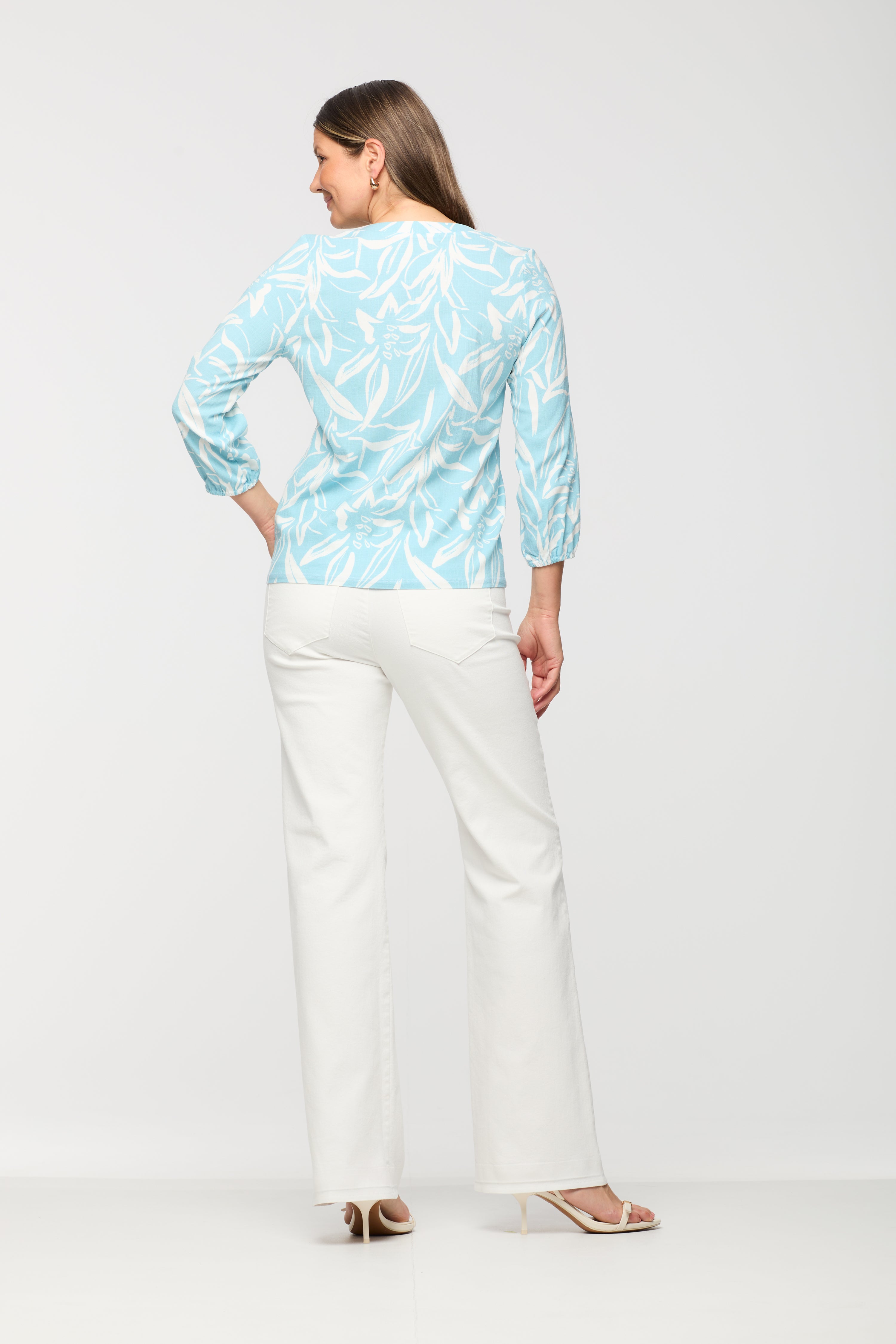 Woman wearing a light blue and white printed long sleeve top shown from the back, featuring a relaxed fit and straight hem, paired with white straight leg pants.