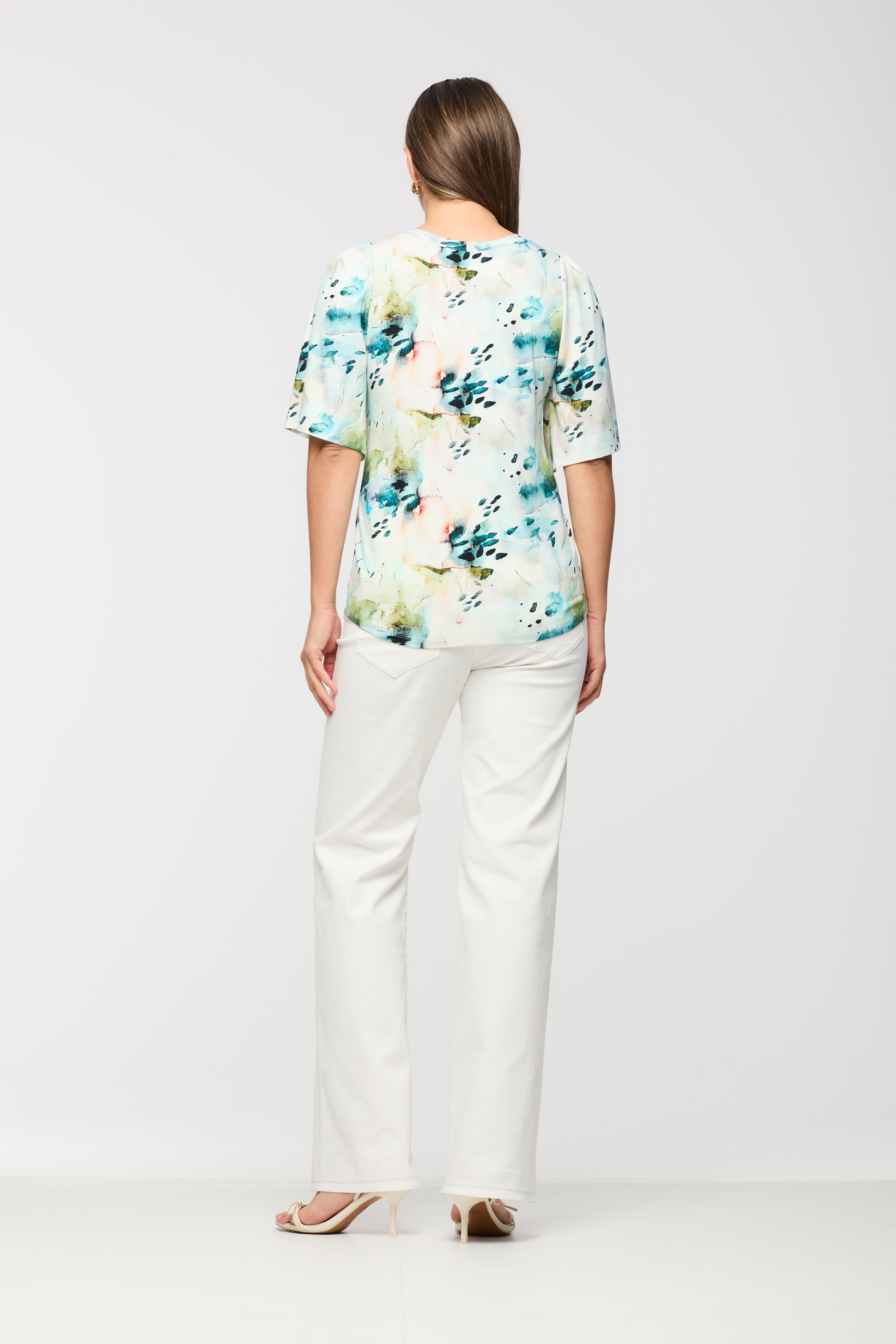Woman wearing a short sleeve printed top shown from the back, featuring a relaxed fit and straight hem, paired with white straight leg pants.