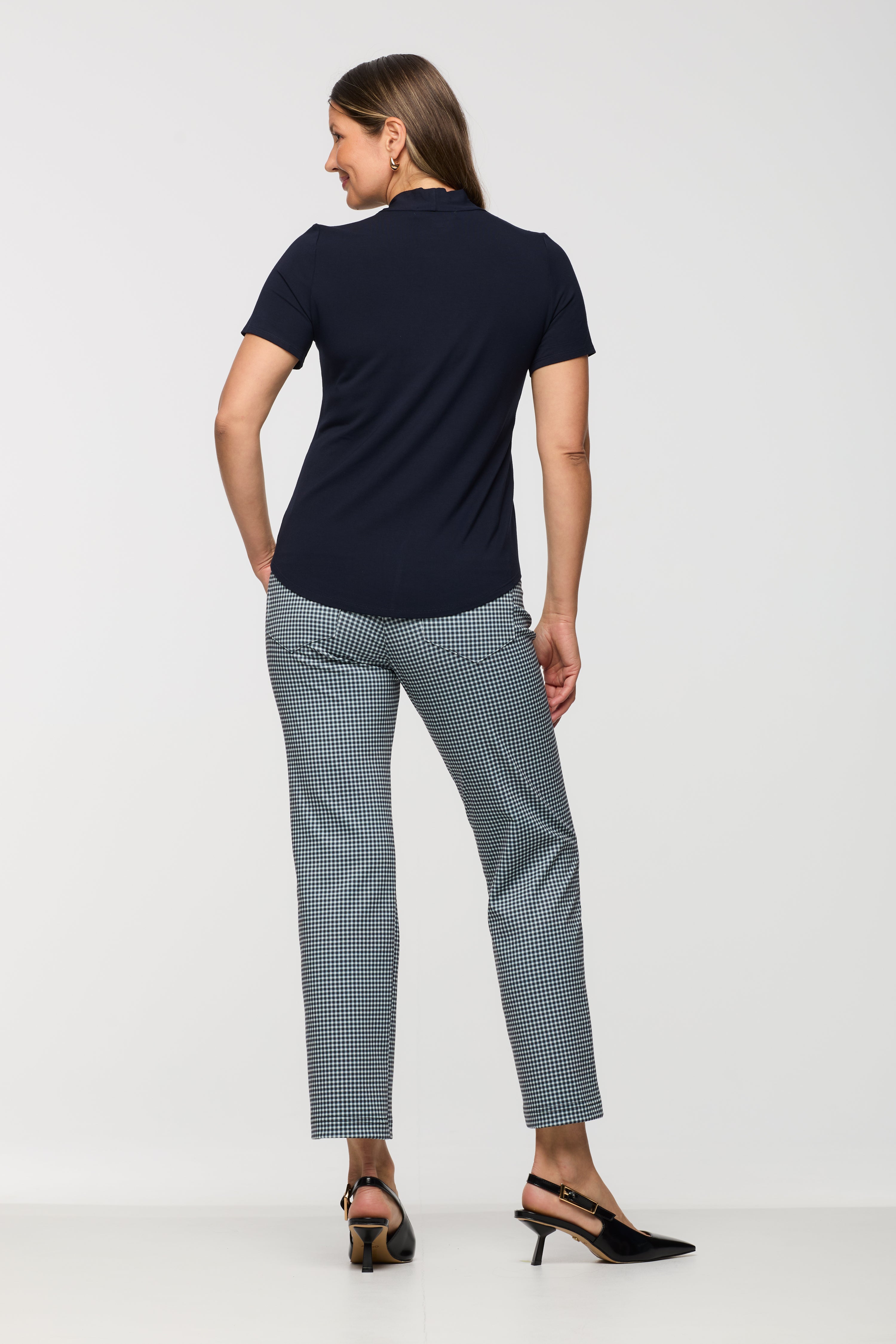 Woman wearing a short sleeve navy top shown from the back, featuring a relaxed fit and curved hem, paired with black and white check cropped straight leg pants.