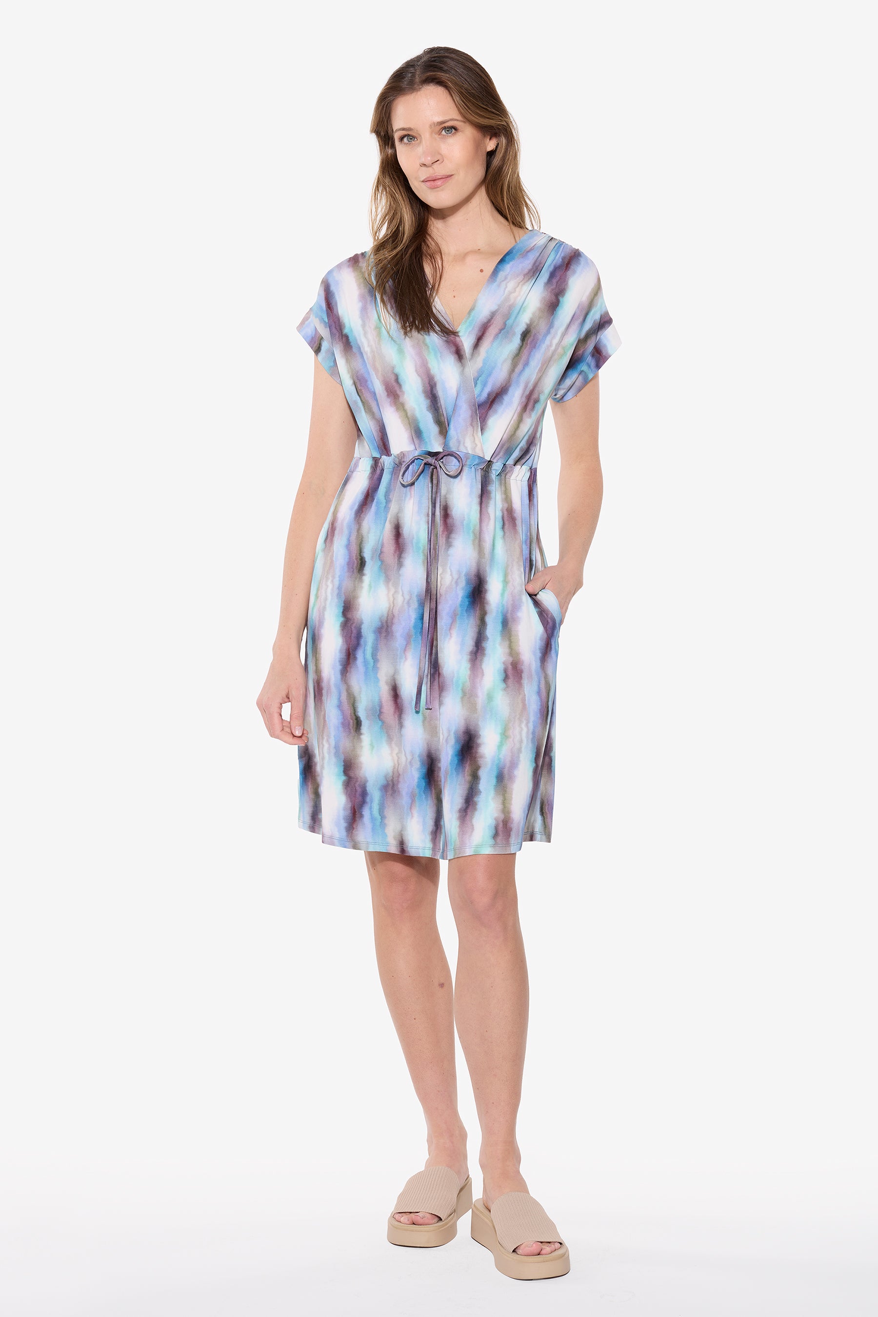 Woman wearing a blue and purple abstract print bamboo dress with short sleeves, V neckline, drawstring waist, and knee-length relaxed fit with hand in pocket