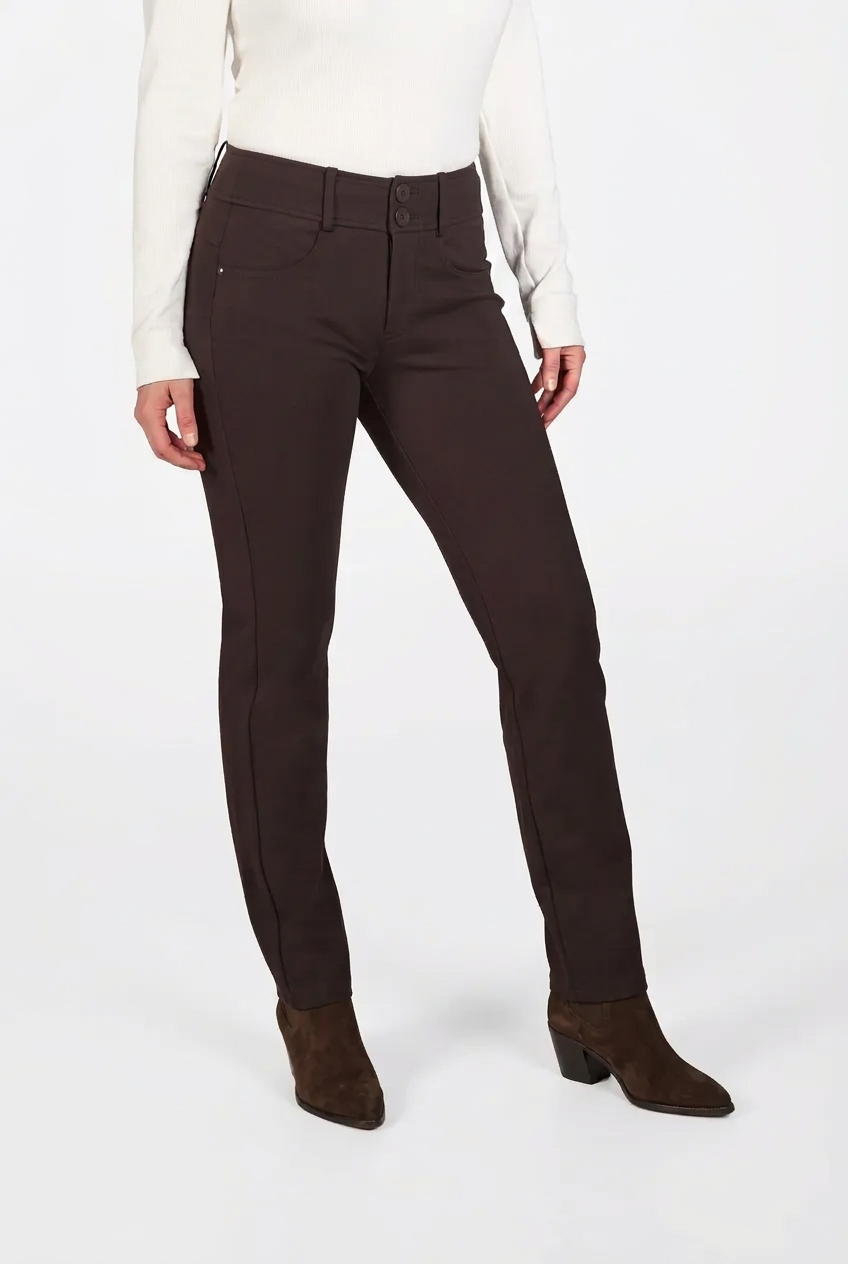 A model wearing a pair of dark brown slim-fit trousers, styled with a white long-sleeve top and brown suede ankle boots, against a plain white studio background.