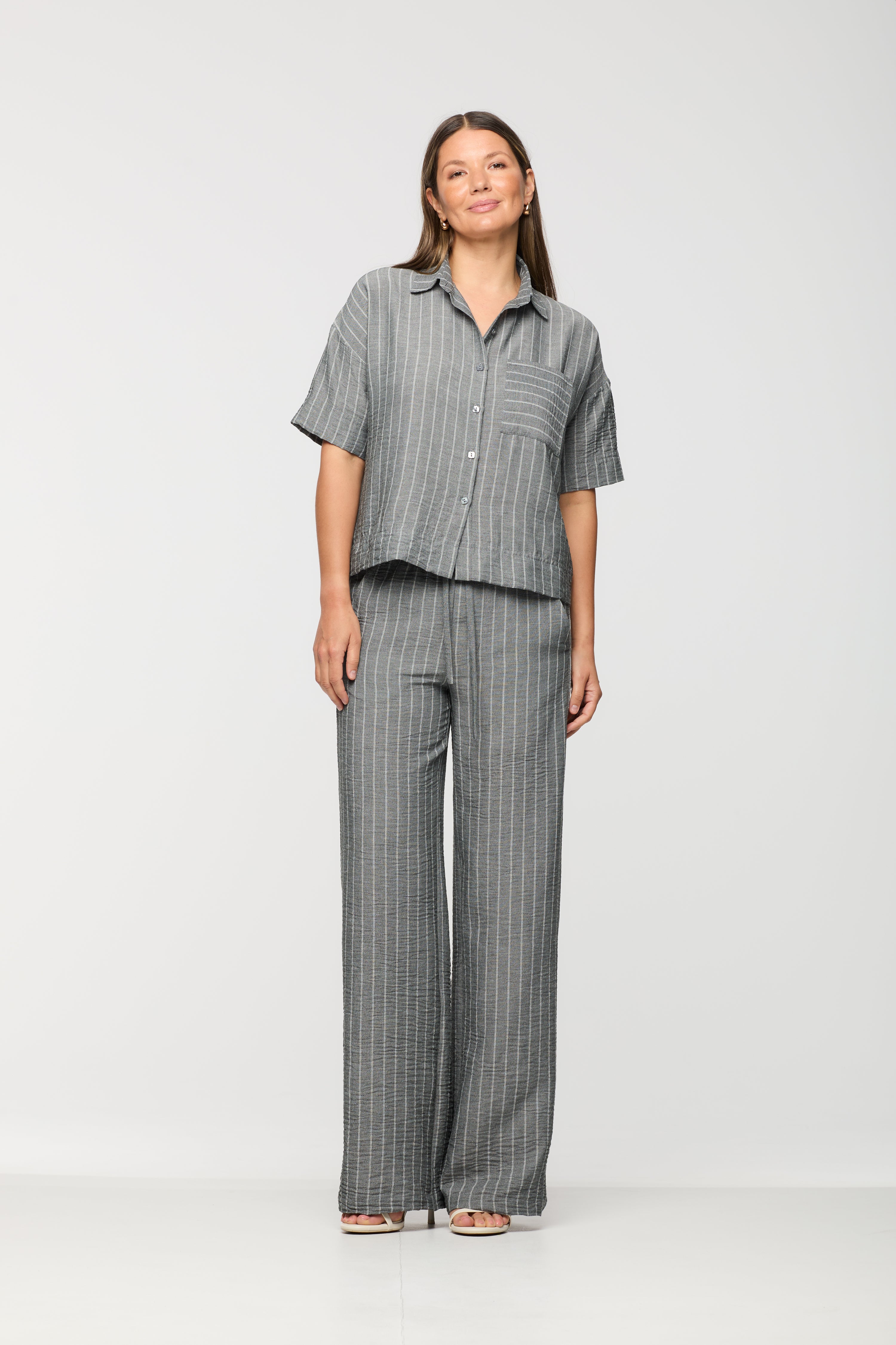 Woman wearing a grey pinstripe short sleeve button-front shirt with a relaxed fit, paired with matching wide leg drawstring pants.