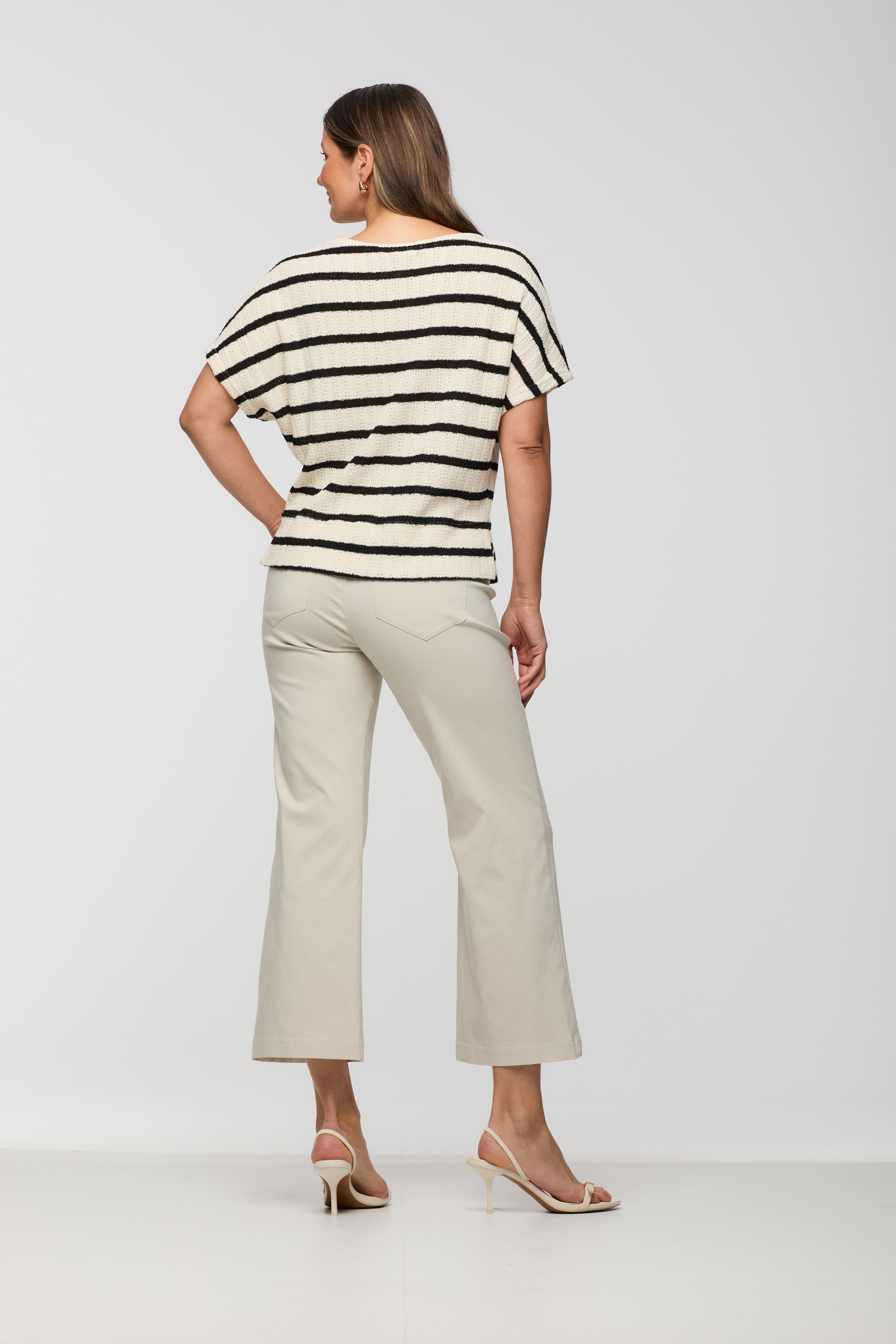 Woman wearing a short sleeve cream textured knit top with black horizontal stripes shown from the back, featuring a relaxed fit, paired with light beige cropped straight leg pants.