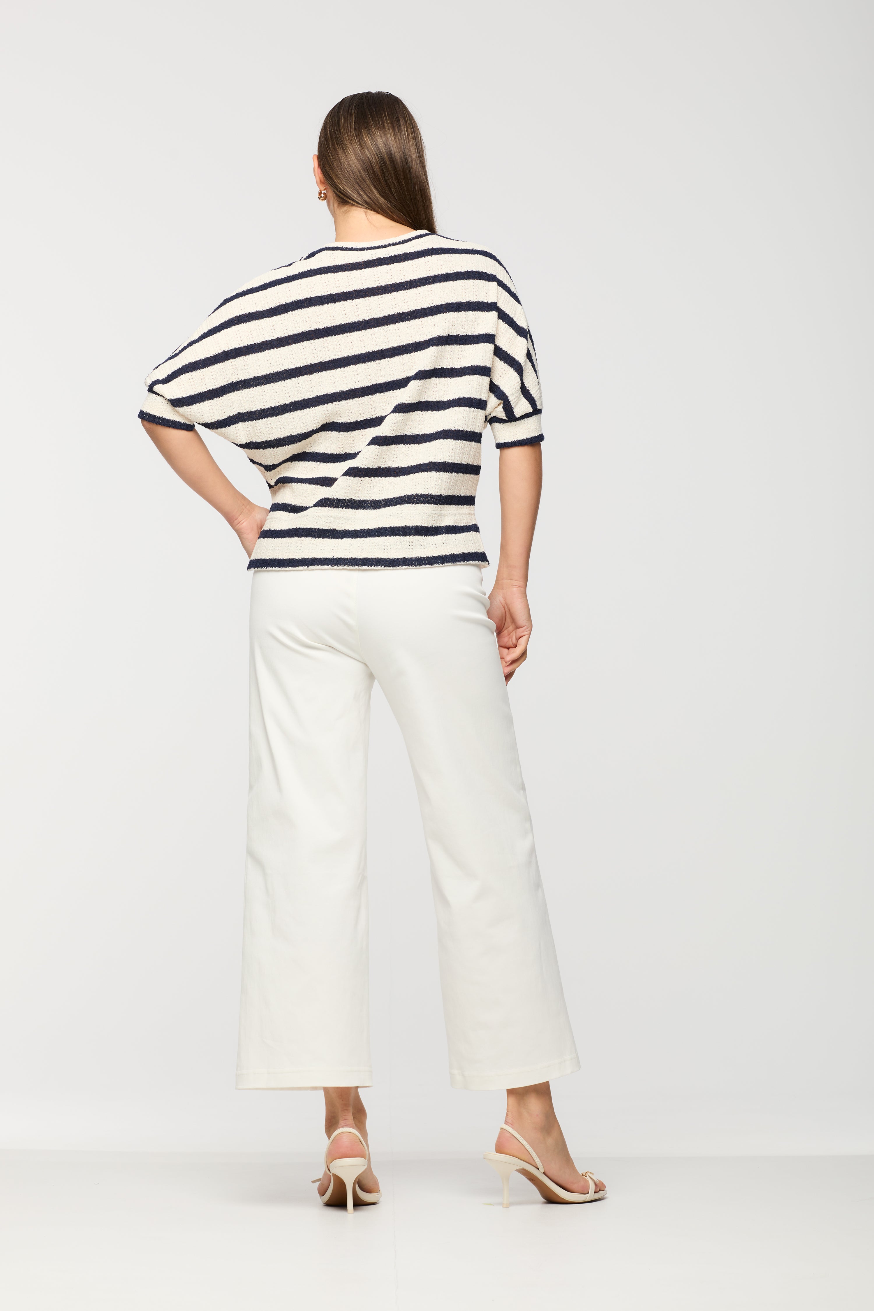 Woman wearing a short sleeve cream knit top with black horizontal stripes shown from the back, featuring a relaxed fit, paired with white wide leg cropped pants.