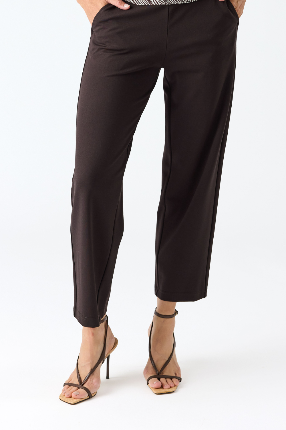 A person wearing dark brown cropped pants with a textured top and strappy high-heeled sandals, shown from the waist down on a light gray studio background.