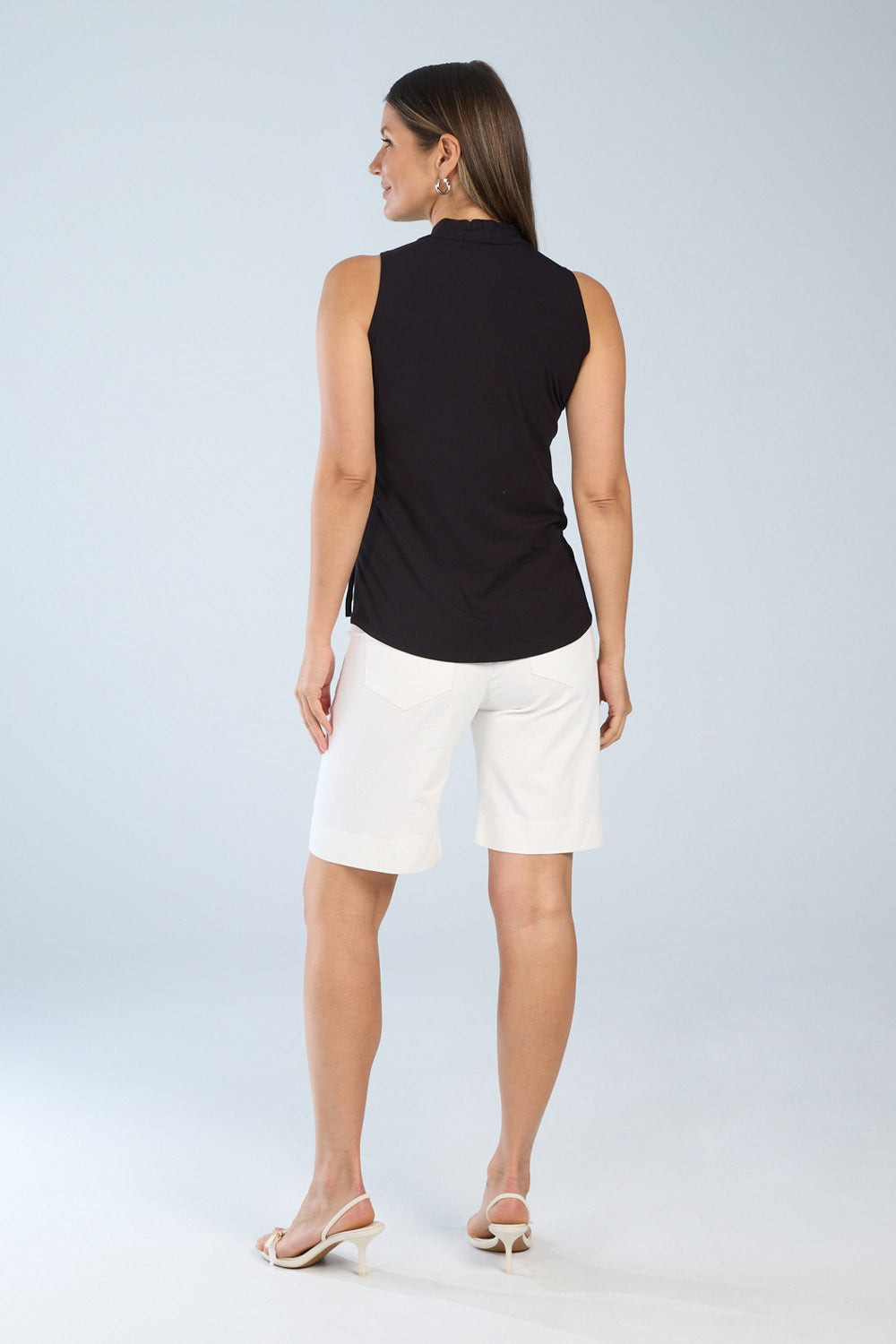 Woman wearing a sleeveless black wrap-style top shown from the back, featuring a relaxed fit and straight hem, paired with white tailored knee-length shorts.