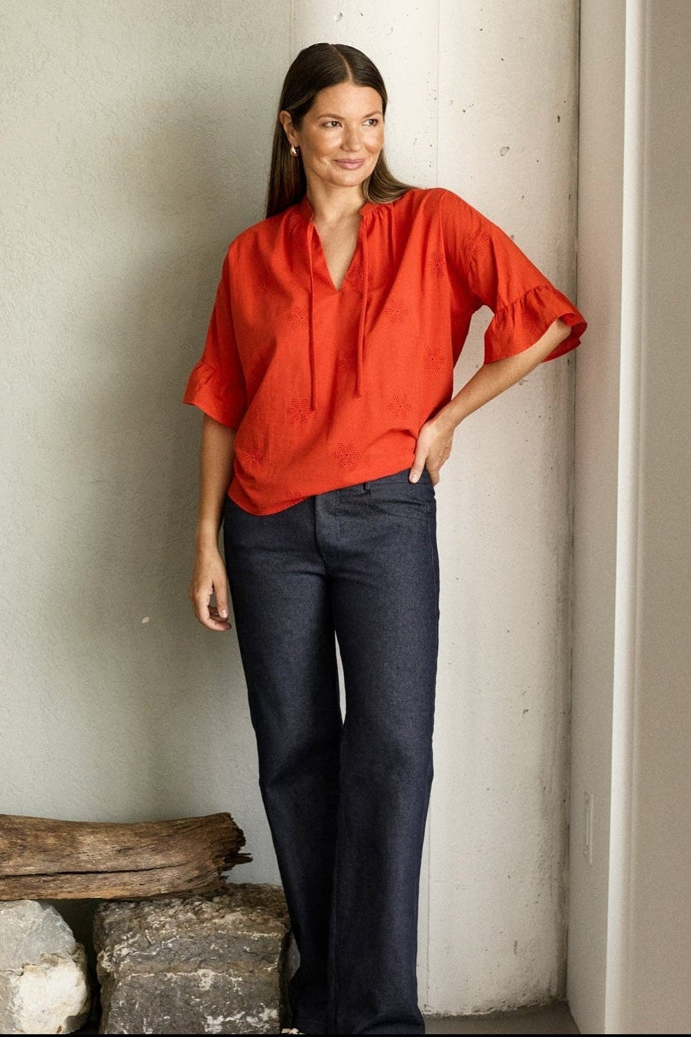Woman in a red blouse and dark pants standing in a minimalistic room with light-colored walls and a window.