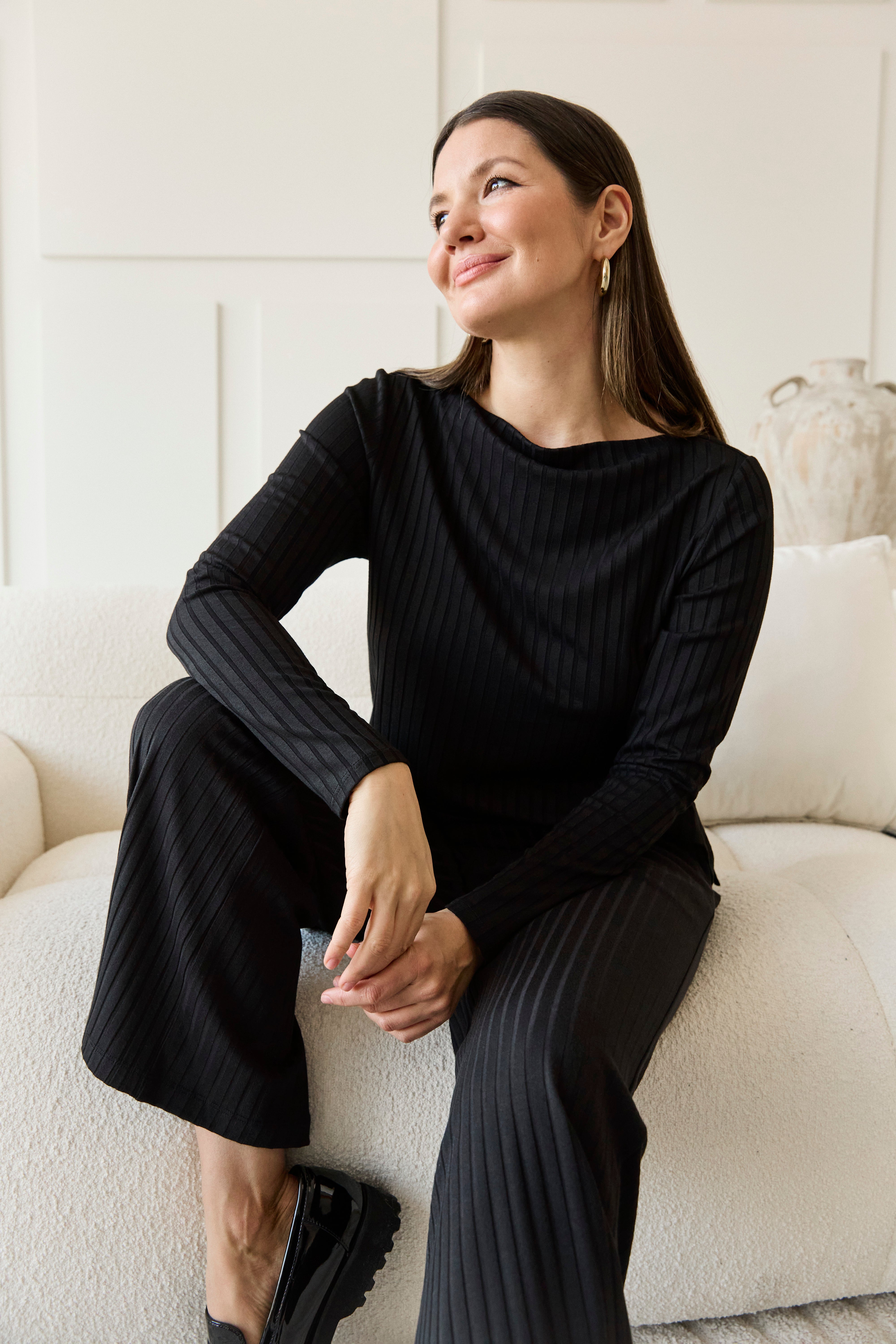 Woman in a black outfit sitting on a white couch in a bright room.