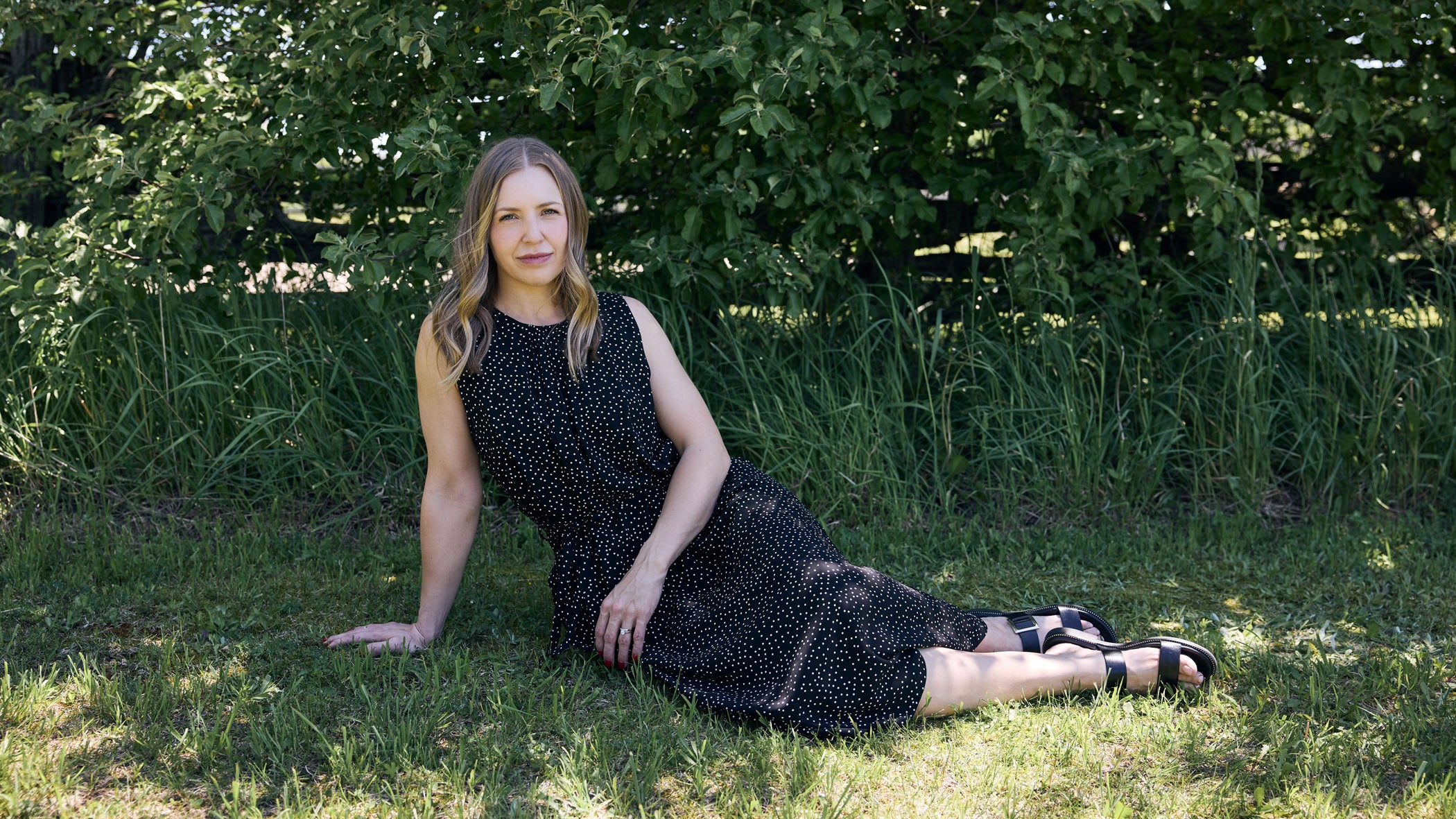 Woman in a black dress sitting on grass with greenery in the background